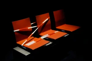Im chiaroscuro Stil sieht man drei leuchtend orange-rote Sitzplätze. Zwei davon wurden während Corona mit einem Band abgesperrt. Farbfoto. (Bastian Peter, Street Photography Schweiz)