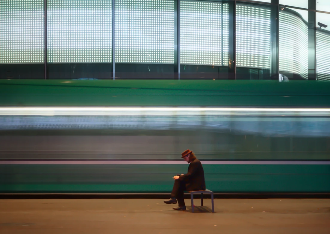 Man sieht einen Mann mit Hut sitzend an der Tramstation Messeplatz in Basel, in der Schweiz. Das Tram rast an ihm vorbei, wobei der Effekt durch Slow Shutter Speed verstärkt wird.