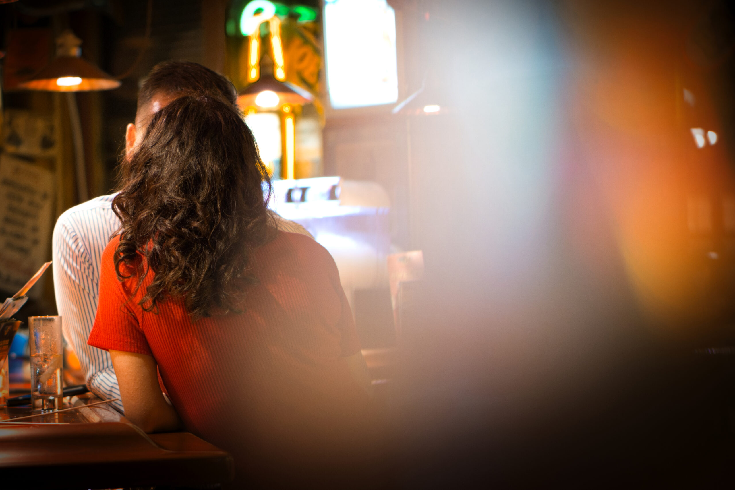 A couple talking intensively at a bar. We do not see faces, the foreground is blurry and the woman is wearing red. Street Photography at night. Photo by Bastian Peter.